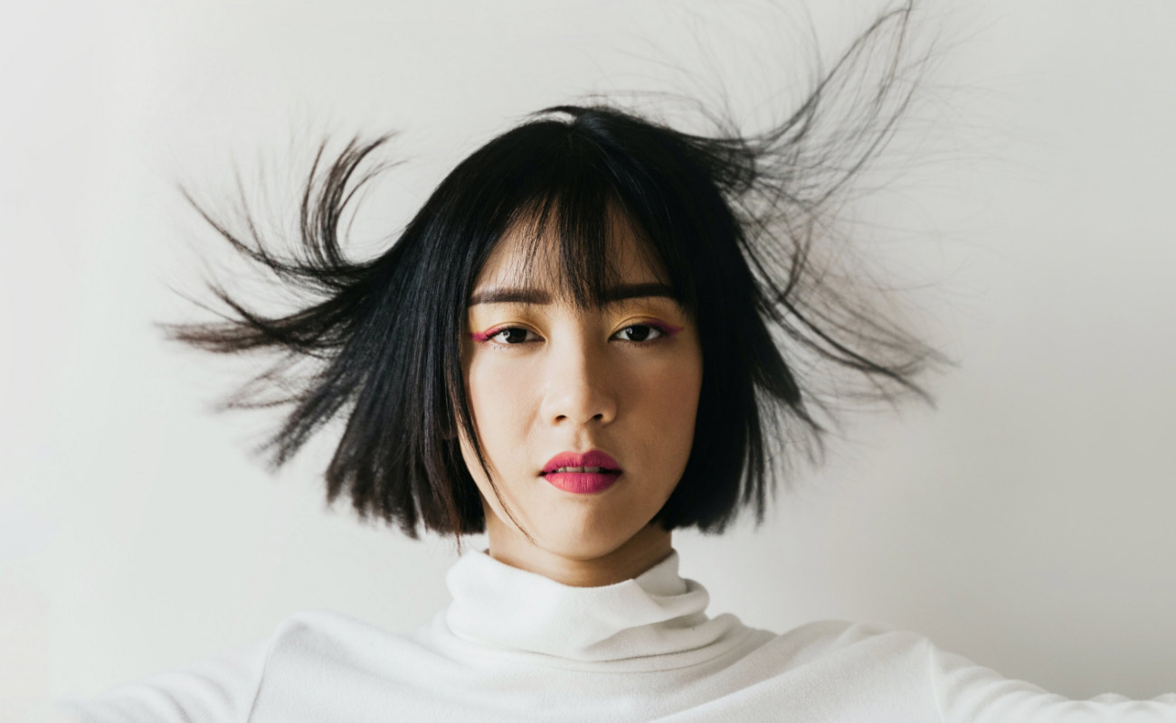 Woman with short black hair styled in a playful manner, wearing a white turtleneck, showcasing vibrant makeup, emphasizing creative hairstyle options like 'Curled Buns.'